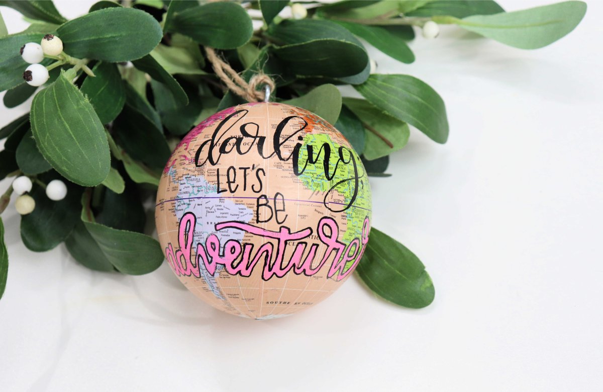 Image contains a peach colored globe ornament with the words, "darling let's be adventurers," and mistletoe.