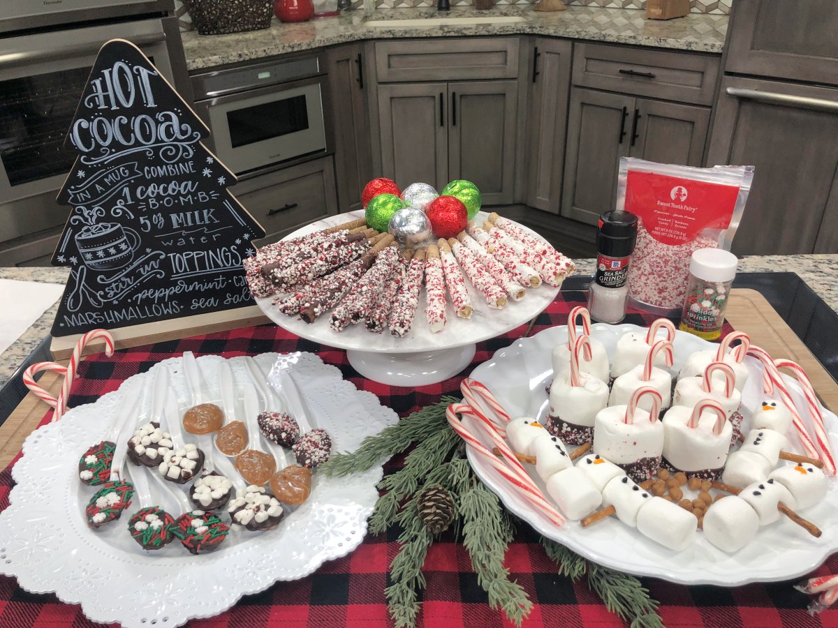 Image contains a hot cocoa bar display with a chalkboard sign, three white plates, and assorted stir-in treats.