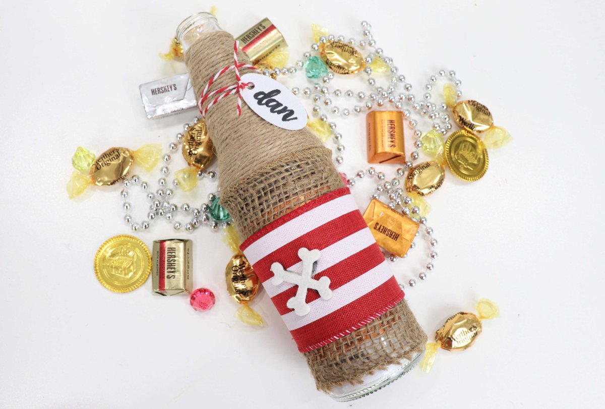 This image contains a jute and burlap wrapped bottle with red and white ribbon, a name tag, and gold wrapped candies.