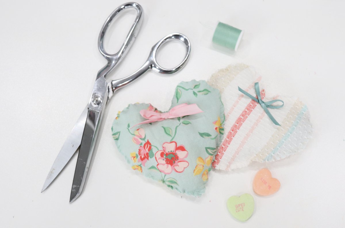 Image contains two fabric hearts; one green with flowers and one with multi-colored stripes, surrounded by scissors, thread, and two candy hearts on a white background.