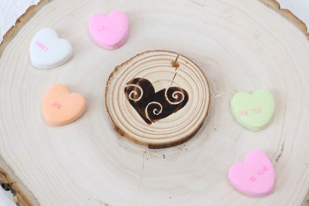 Image contains a small wood slice with a wood burned heart design on top of a larger wood slice decorated with candy hearts.