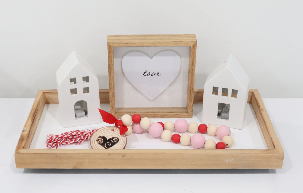 Image contains a decorative tray styled with two white ceramic houses, a wooden bead garland, and a sign with a heart that reads, "love."