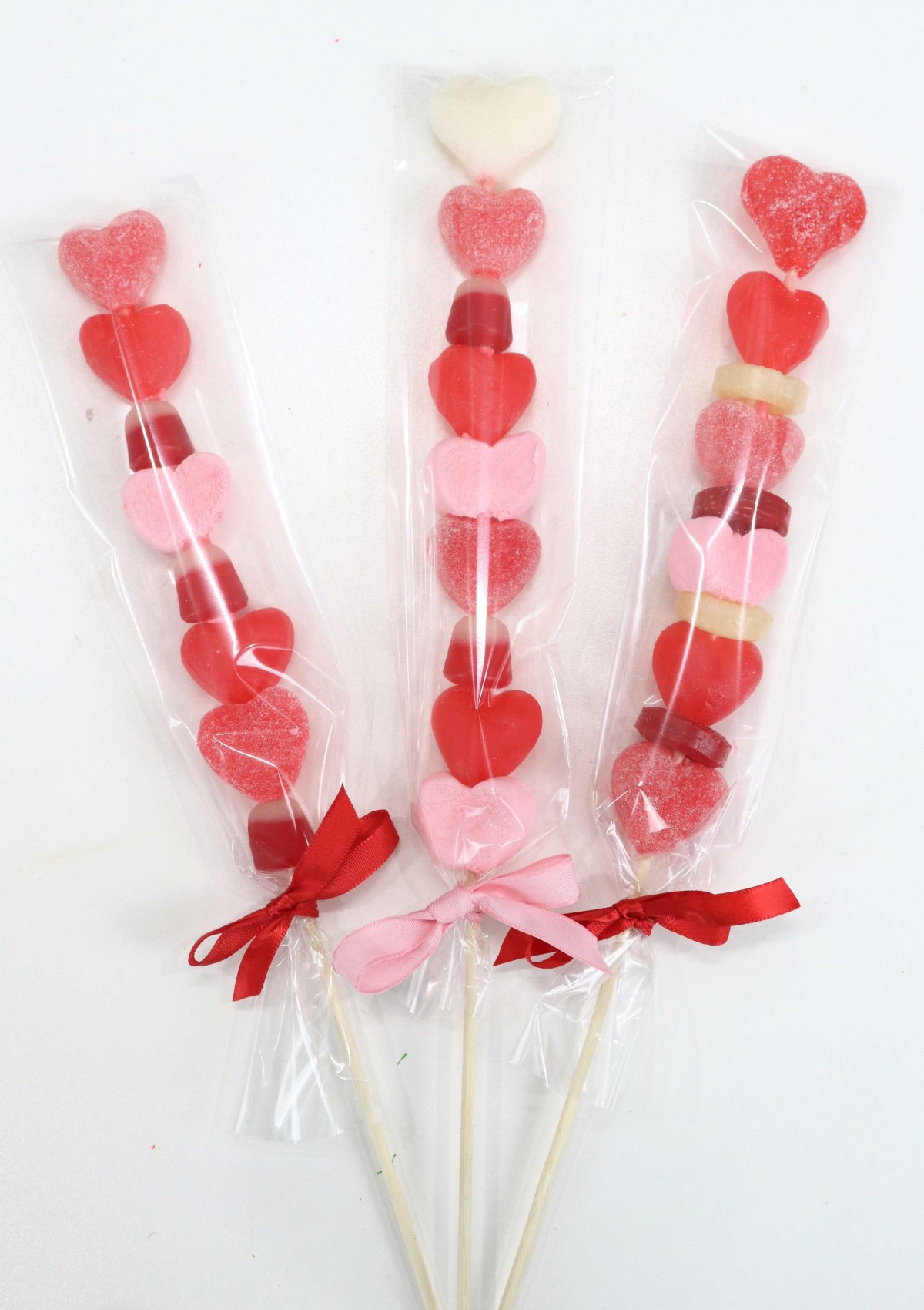 Image contains three candy kabobs with gummy heart candies, marshmallows, and other red, pink, and white candies.
