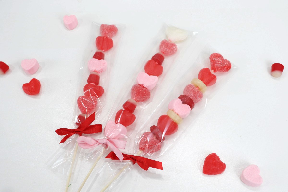 Image contains assorted heart shaped red and pink candies on skewers with more candy scattered on a white table.