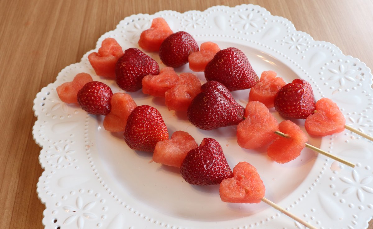 Image contains three fruit kabobs; heart shaped watermelon and strawberries on skewers, on a white plate.