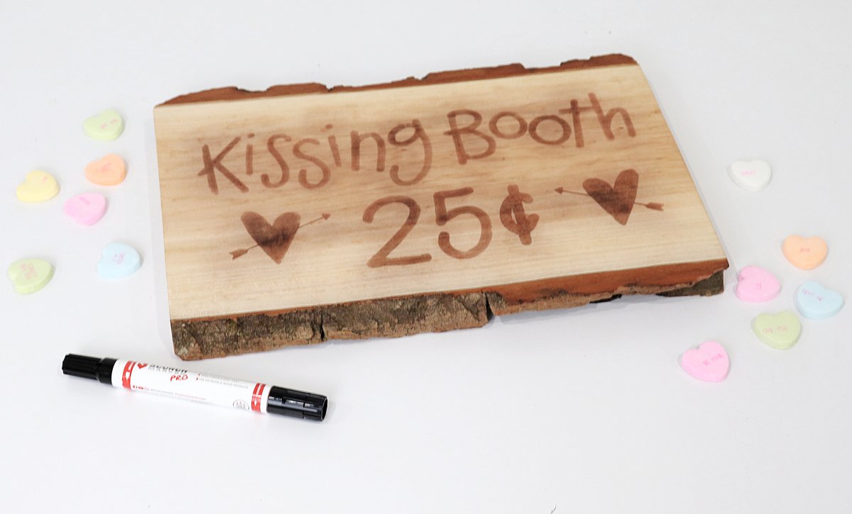Image contains a wooden sign that reads, "kissing booth 25 cents" with two hearts, along with scattered candy hearts and a Scorch Marker.