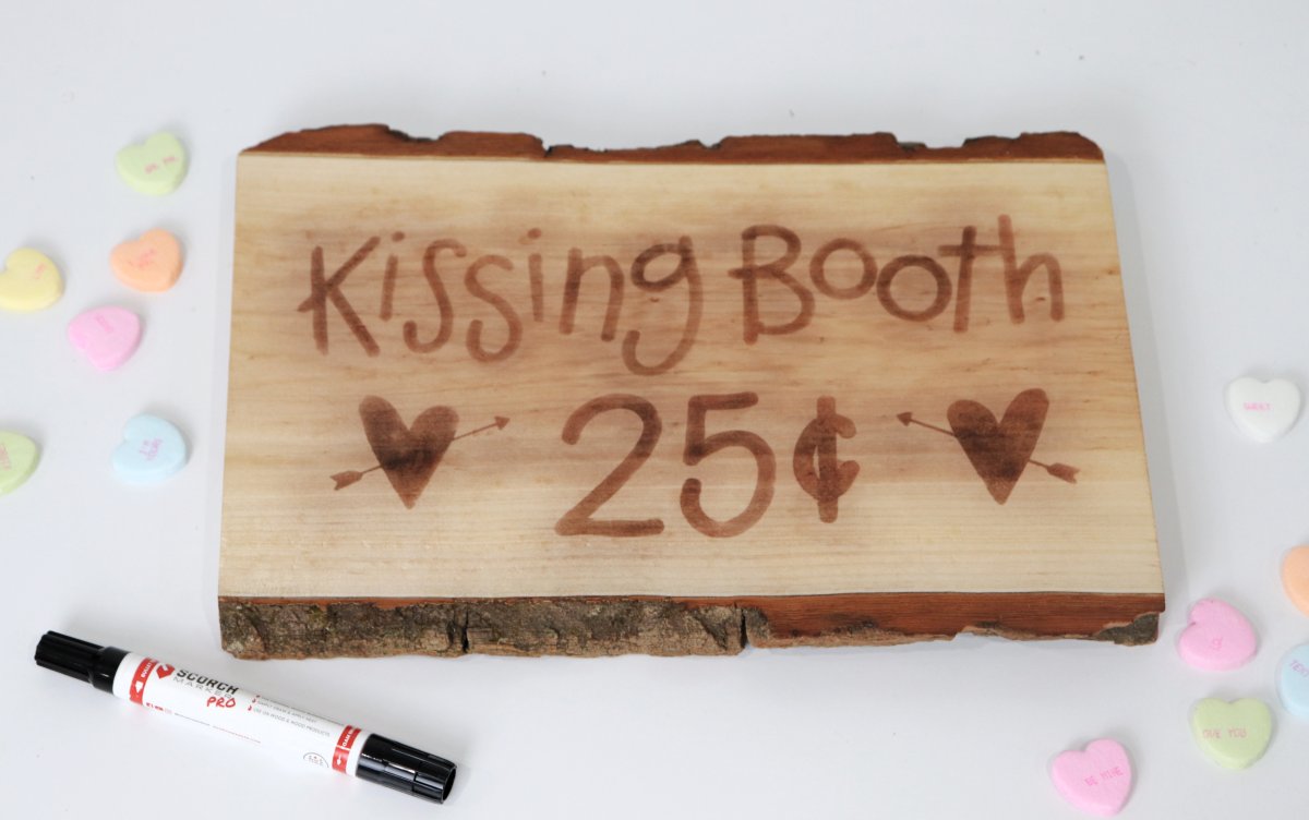 Image contains a wooden sign that reads, "kissing booth 25 cents" and has two hearts, on a white background with scattered candy hearts and a Scorch Marker.