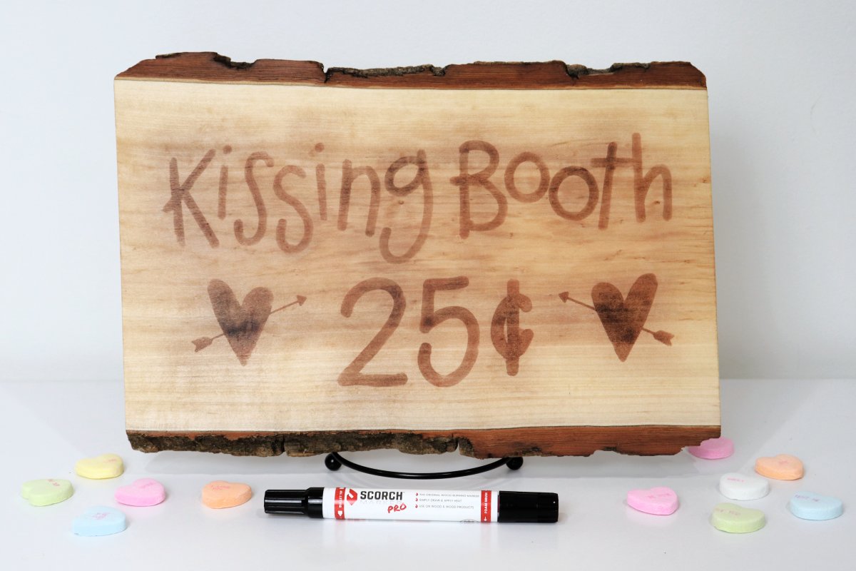 Image contains a wooden sign with the words "kissing booth, 25 cents) and two hearts, on a white background with candy hearts and a marker.