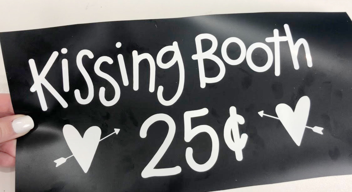 Image contains a hand holding a black stencil with the words "kissing booth 25 cents" and two hearts with arrows.