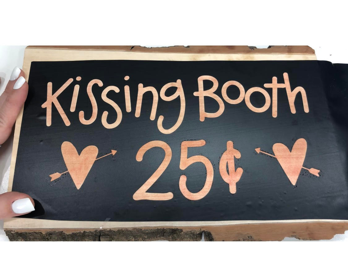 Image contains a black stencil on a piece of wood that reads, "kissing booth 25 cents" and has two hearts with arrows.