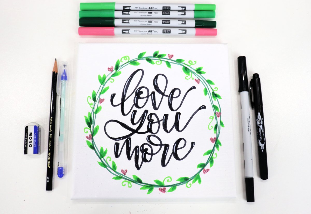 Image contains a hand lettered canvas with the words "love you more," surrounded by markers, a pencil, a glue pen, and an eraser.