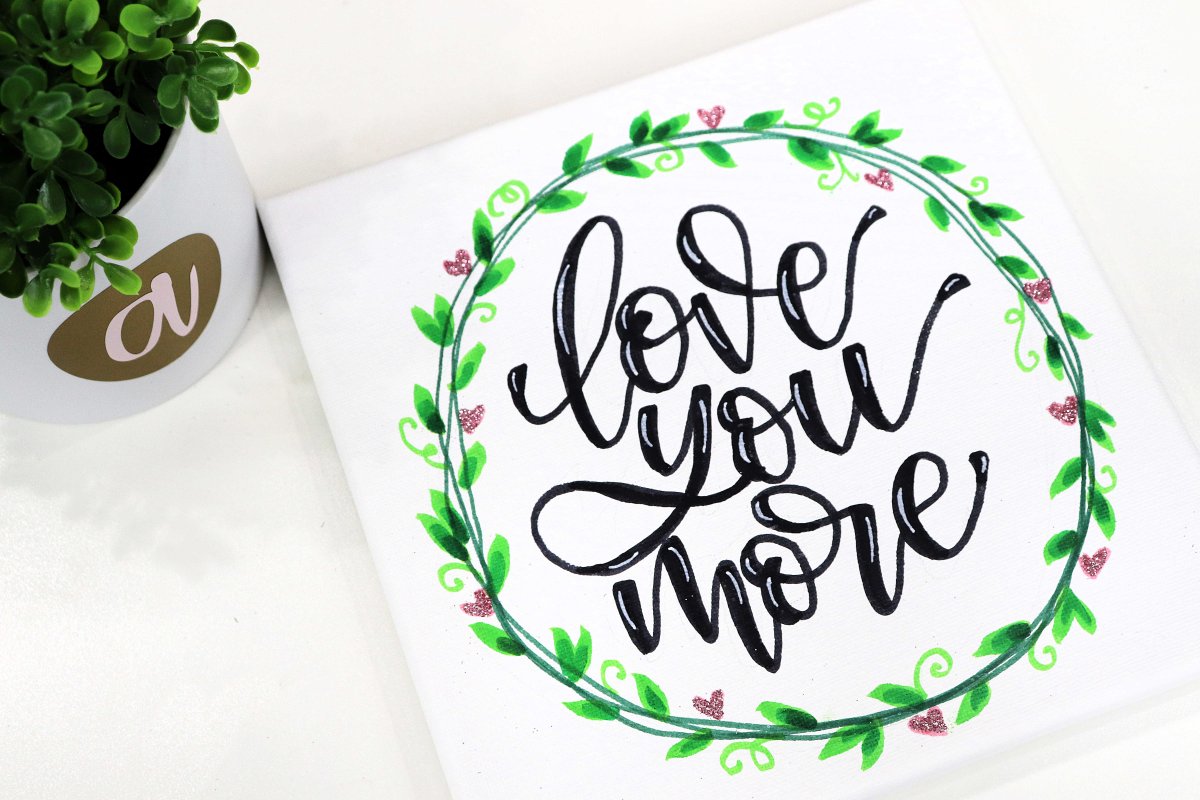 Image contains a square canvas with a wreath and the words, "love you more," next to a plant.