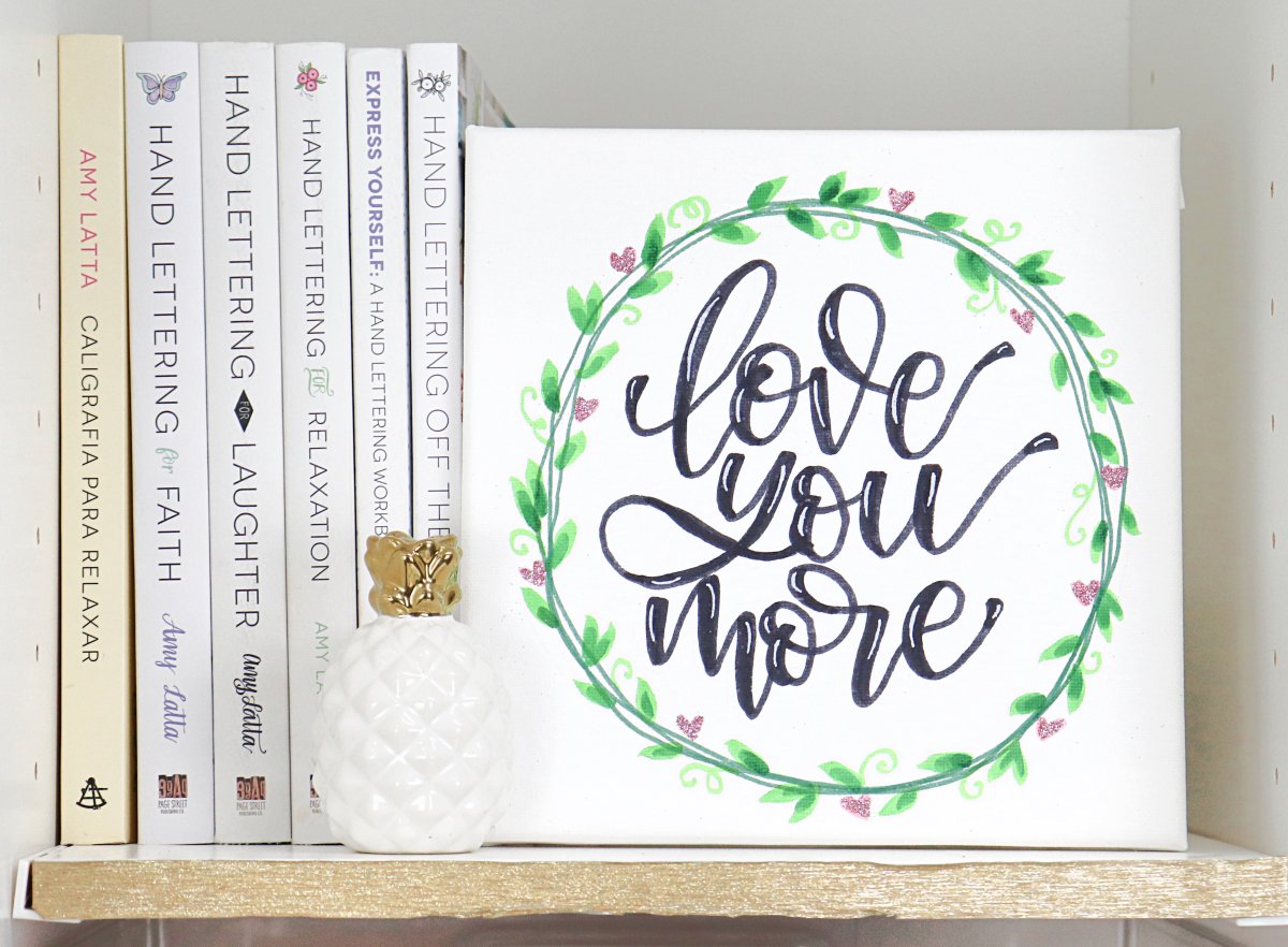 Image contains a shelf with multiple books, a decorative pineapple, and a canvas with the words, "love you more."