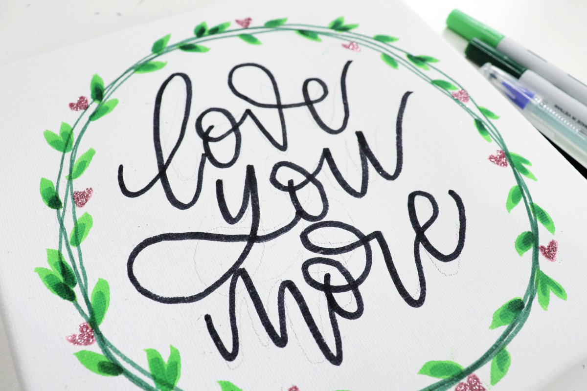 Image contains a canvas with an illustrated wreath and the words, "love you more" along with several markers.