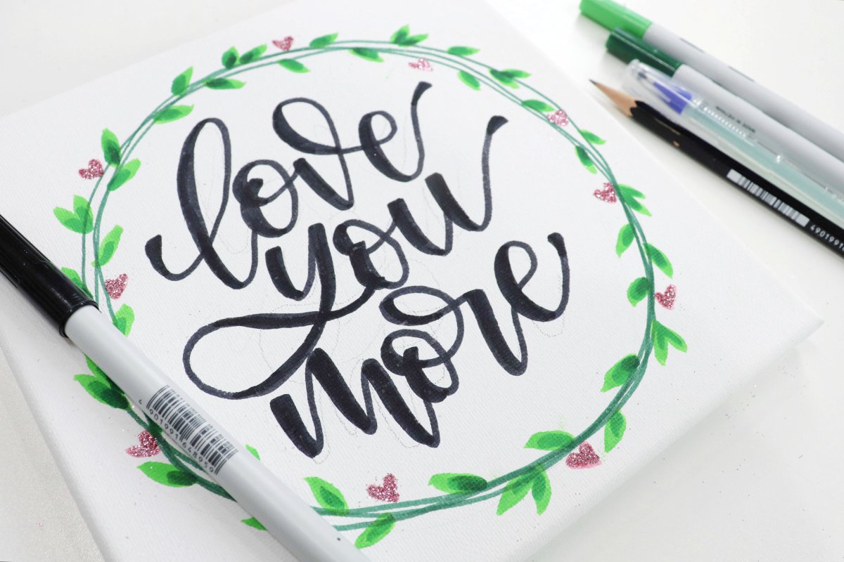 Image contains a hand lettered canvas that reads, "love you more," along with assorted markers and a pencil.
