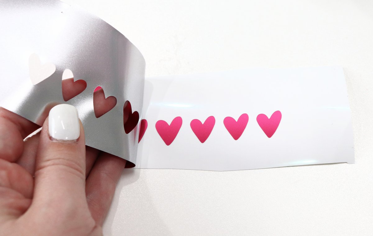Image contains a hand peeling vinyl away to reveal a series of small pink vinyl hearts.