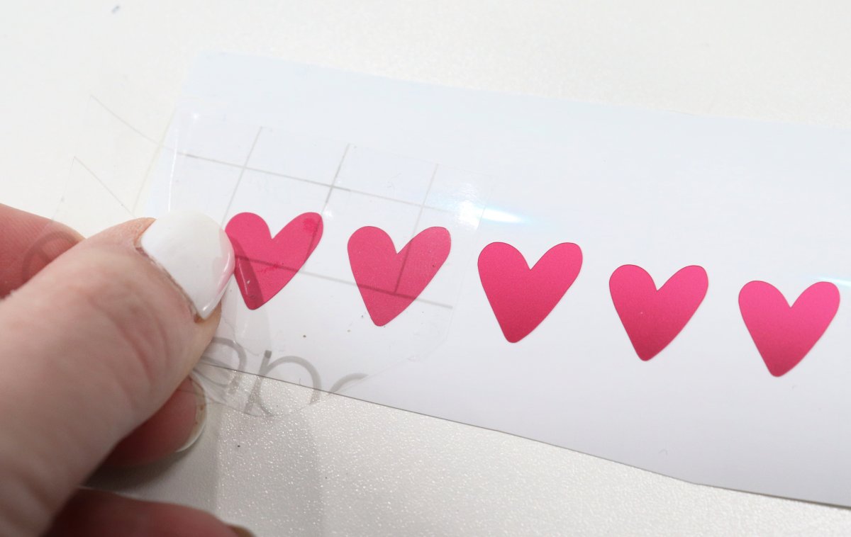Image contains a hand applying a piece of clear transfer paper to a row of pink vinyl heart cut-outs.