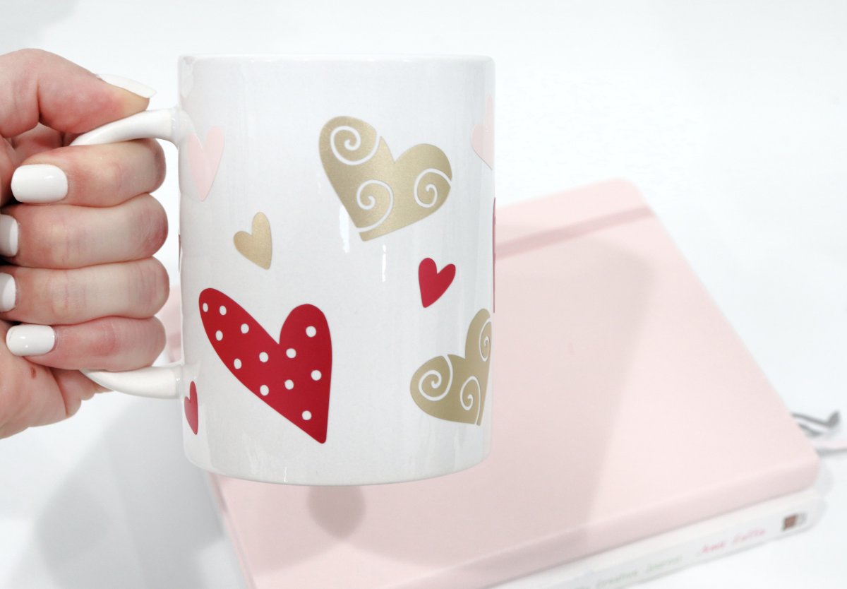 Image contains a hand holding a white coffee mug decorated with hearts in shades of pink and gold, in front of a stack of pink and white books.