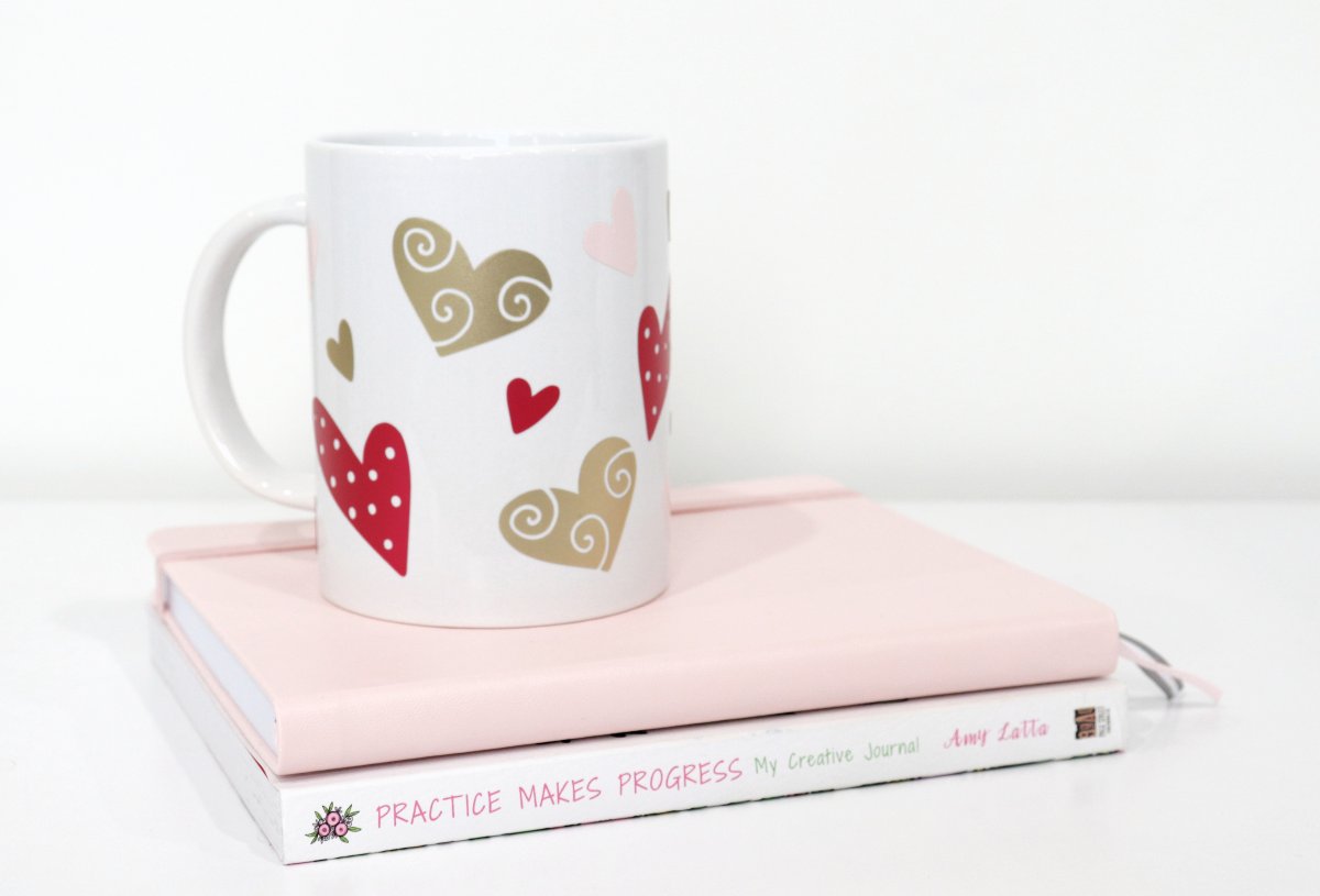 Image contains a white coffee mug covered with pink and gold hearts, sitting on top of a white and pink book stack.