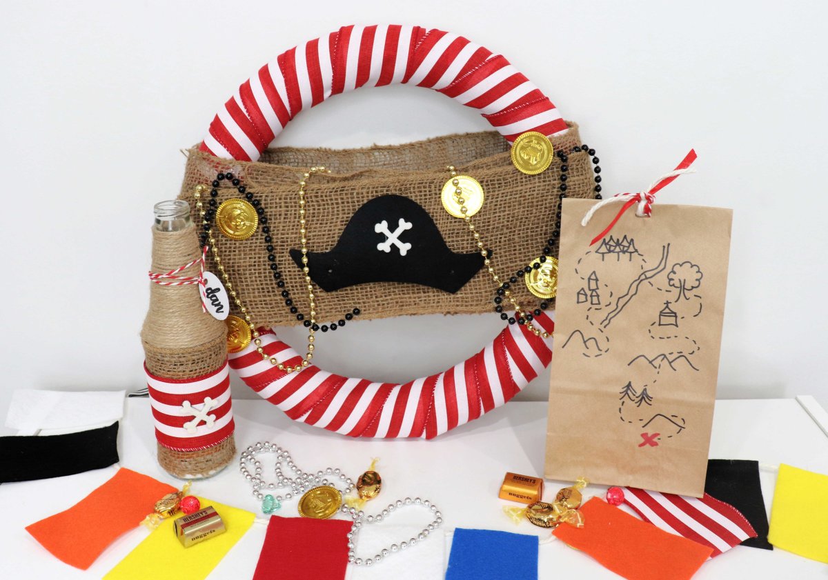 This image contains a pirate themed wreath, a message in a bottle invitation, a paper bag party favor, a colorful felt flag banner, and candy.