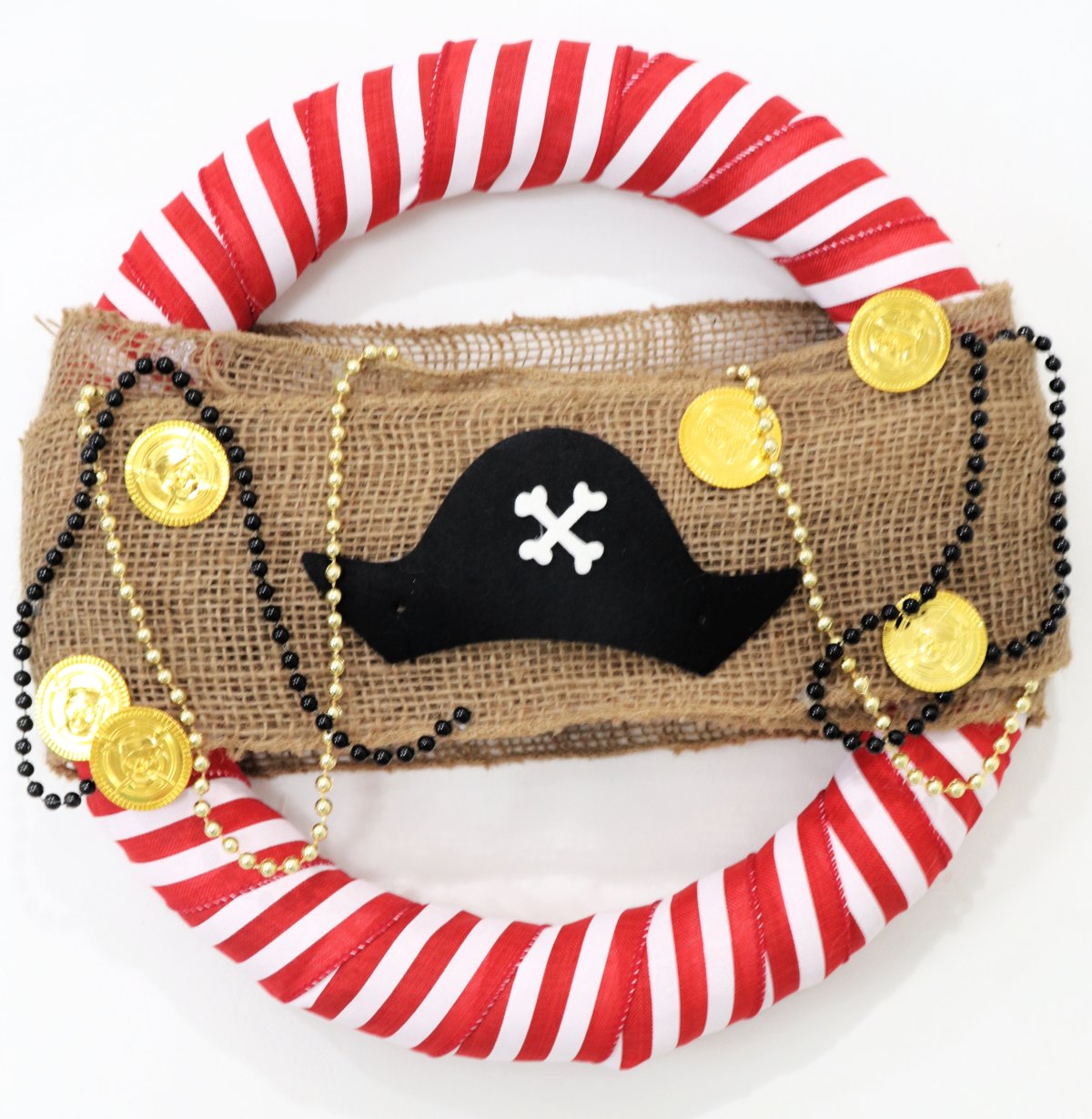 This image contains a wreath wrapped in red and white striped ribbon with burlap across the center, a pirate hat, beads, and gold coins.