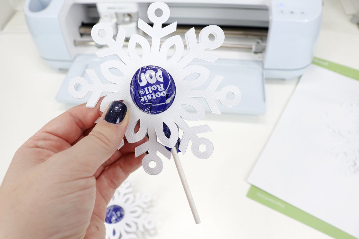 Image contains a hand holding a decorated snowflake lollipop, a Cricut machine, and a cutting mat.