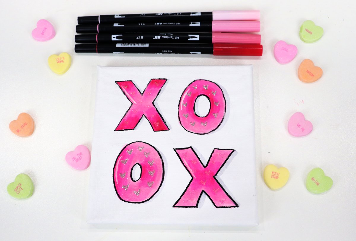 Image contains four pink markers, candy hearts, and a square canvas with pink "xo" letters.