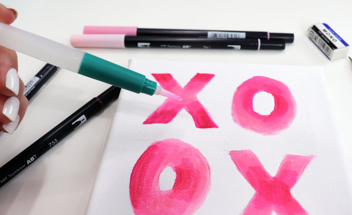 Image contains assorted markers and a canvas with pink "xo" letters, along with a water pen.