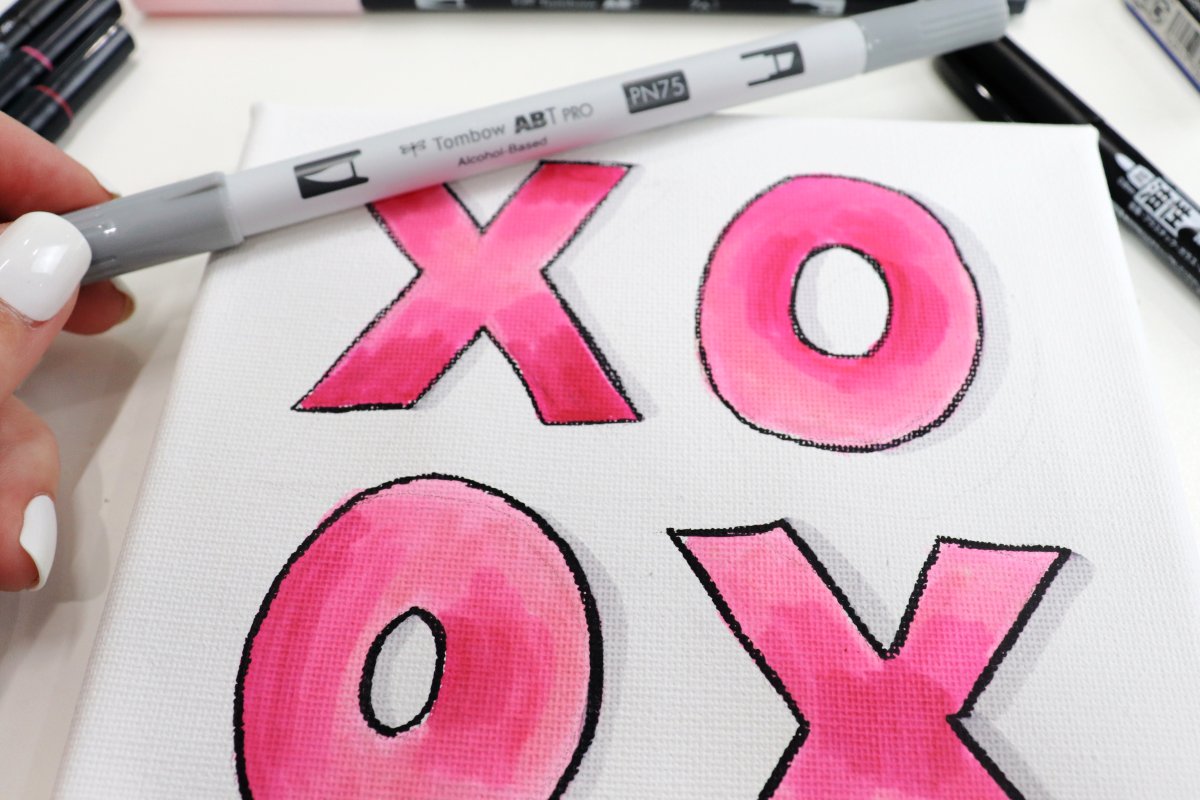 Image contains a hand holding a grey marker and a square canvas with pink "xo" letters.
