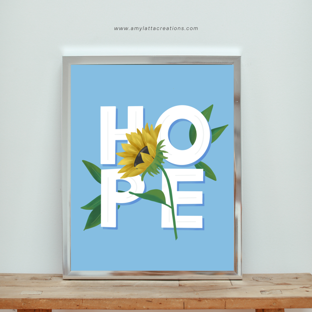 Image contains a printable with the word HOPE in white letters on a blue background with a sunflower and green leaves. The printable is in a silver frame, sitting on a wooden table against a grey wall.
