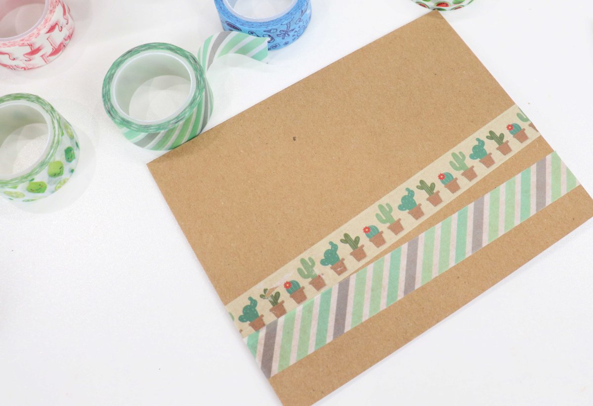 Image contains a brown handmade card with two stripes of washi tape across the front. It sits on a white background surrounded by rolls of assorted colors of washi tape.