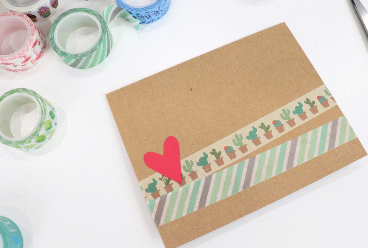 Image contains a brown handmade card with two strips of washi tape across the front, and a red heart. It sits on a white background surrounded by multi-colored rolls of washi tape.