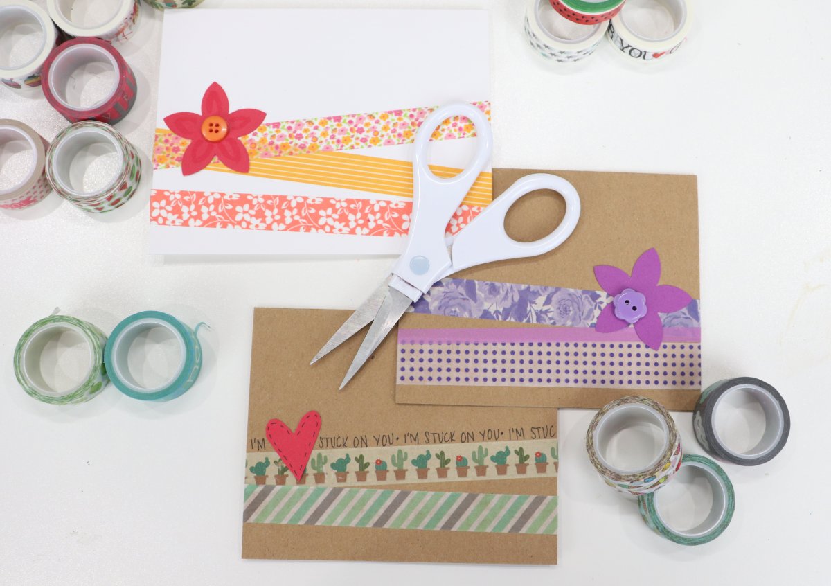 Image contains three handmade cards; two brown and one white, decorated with multi-colored washi tape and cardstock embellishments. They are on a white background, surrounded by rolls of washi tape. A pair of white scissors sits on top.