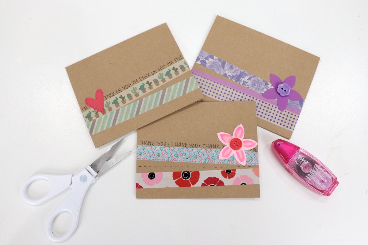 Image contains three kraft paper cards decorated with washi tape and cardstock embellishments. They are on a white background, with a pair of white scissors and a pink adhesive runner.