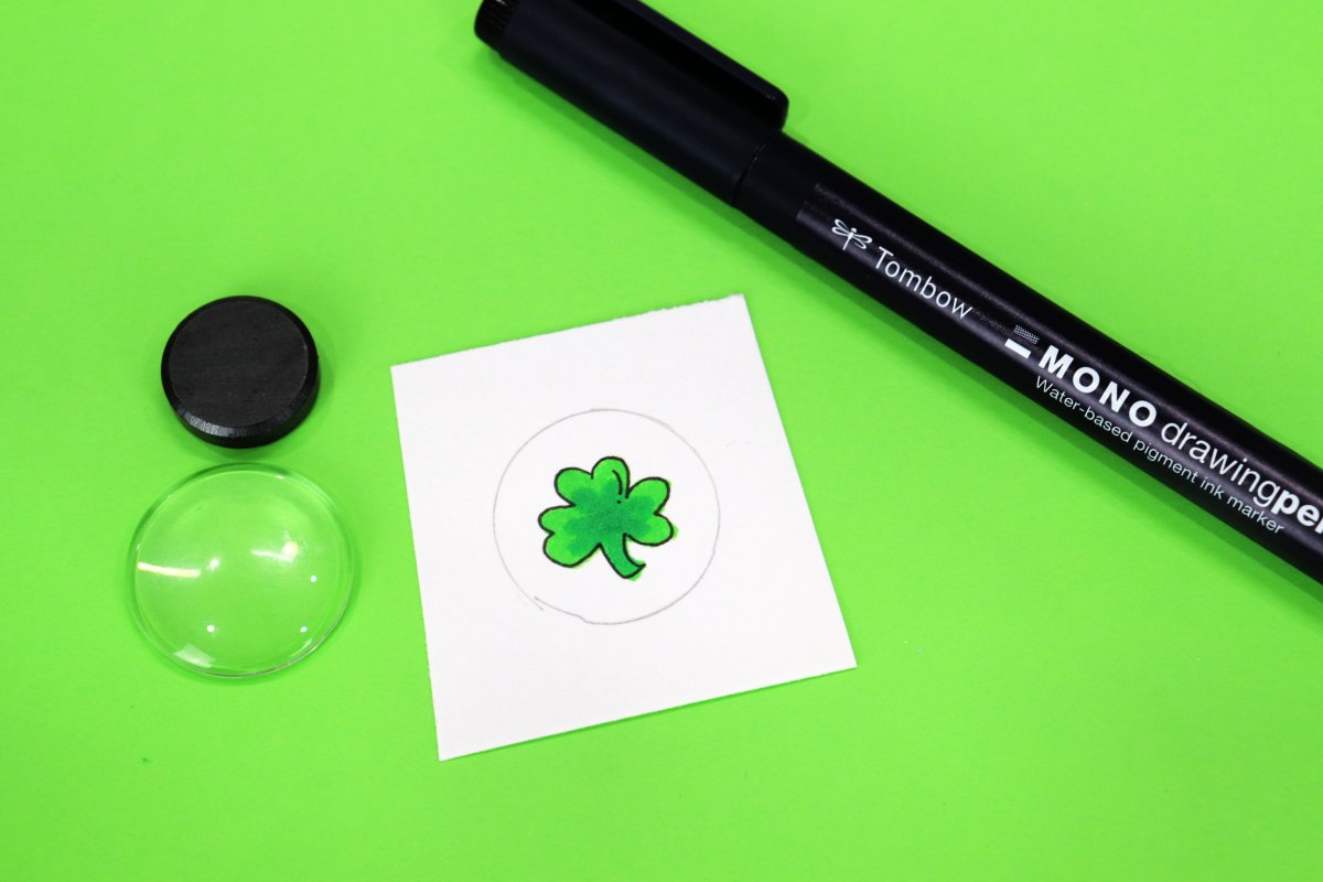 Image contains a white square of paper with a pencil drawn circle and a green shamrock inside. It sits on a green background next to a clear round cabochon, a round magnet, and a black drawing pen.