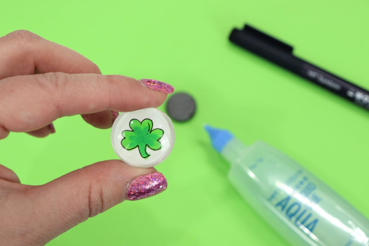 Image contains a hand holding a glass cabochon with a hand-drawn shamrock image glued to the back. This is above a green background that includes the following out-of-focus supplies: a magnet, a tube of clear glue, and a black marker.