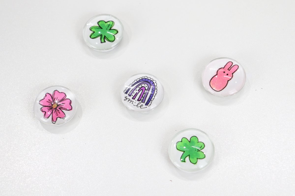 Image contains five round magnets featuring different hand-drawn images. There are two with green shamrocks, one with a pink bunny, one with a pink five-petaled flower, and one with a purple-hued rainbow and the word, "smile."