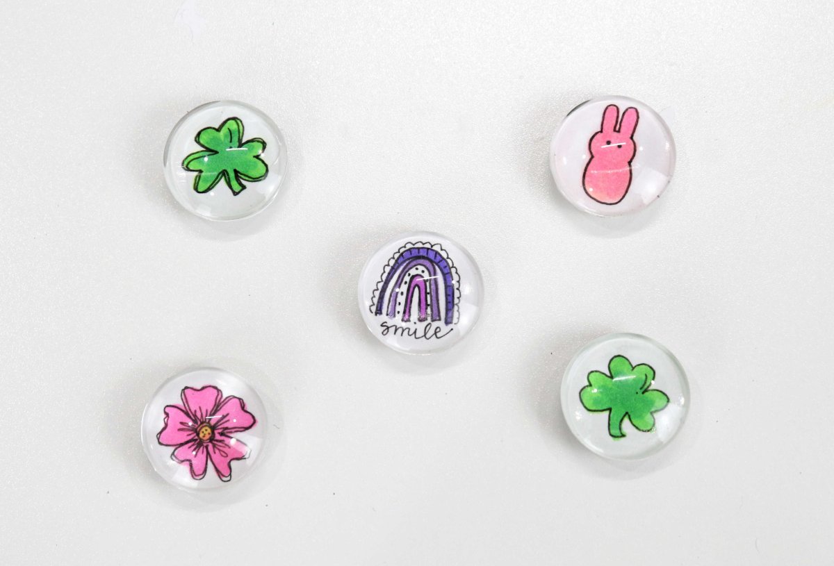 Image contains five round magnets featuring different hand-drawn images. There are two with green shamrocks, one with a pink bunny, one with a pink five-petaled flower, and one with a purple-hued rainbow and the word, "smile."