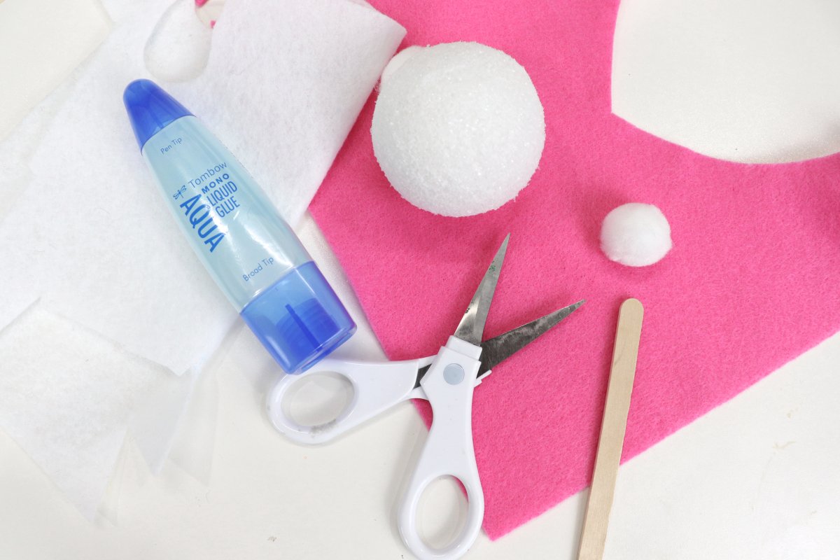 Image contains white and pink felt, white-handled scissors, a small white pom pom, a tube of liquid glue, and a wooden craft stick on a white background.