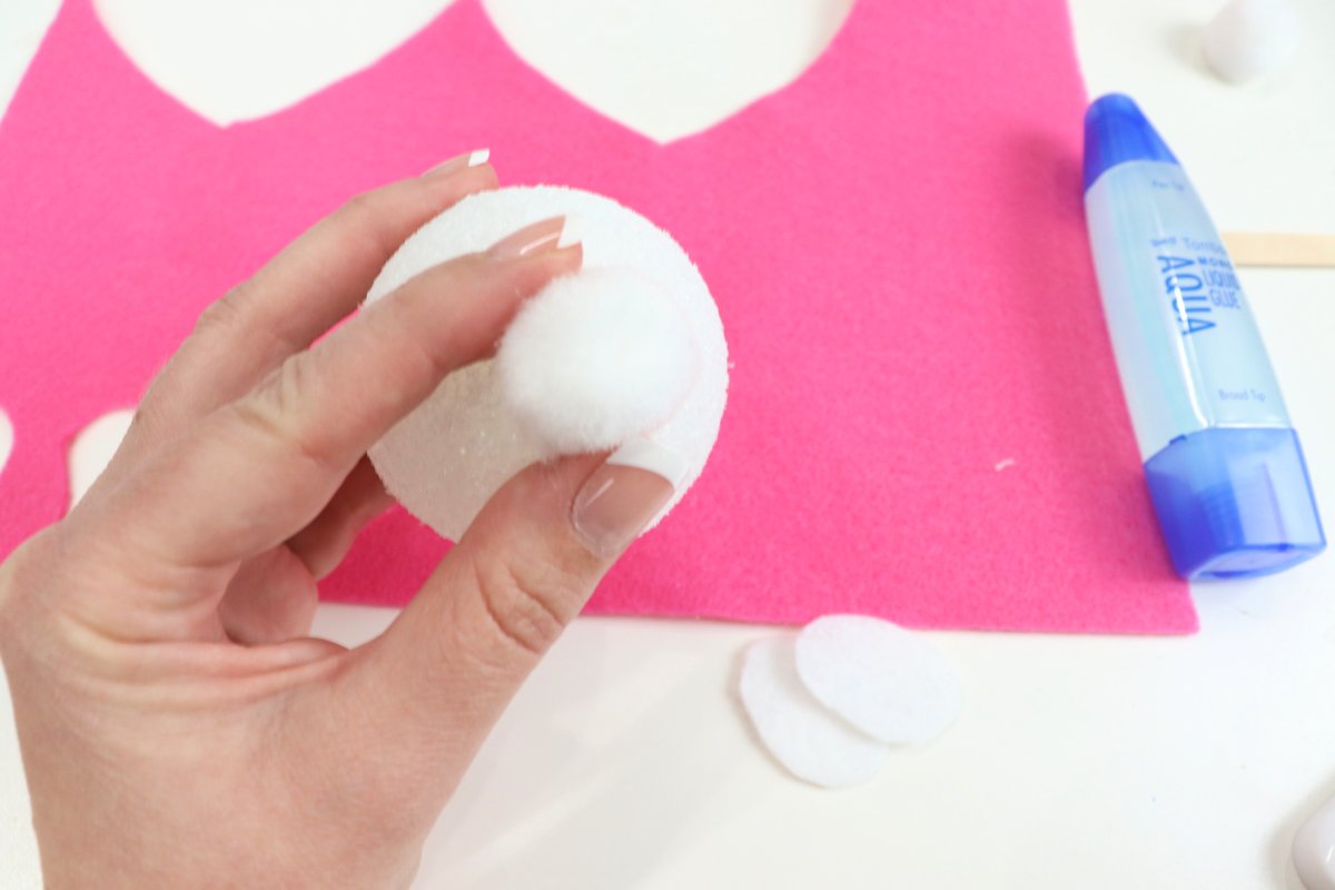 Image contains Amy's hand holding a white pom pom on top of a styrofoam ball. In the background, there is a piece of pink felt, a tube of glue, and some cut white felt.