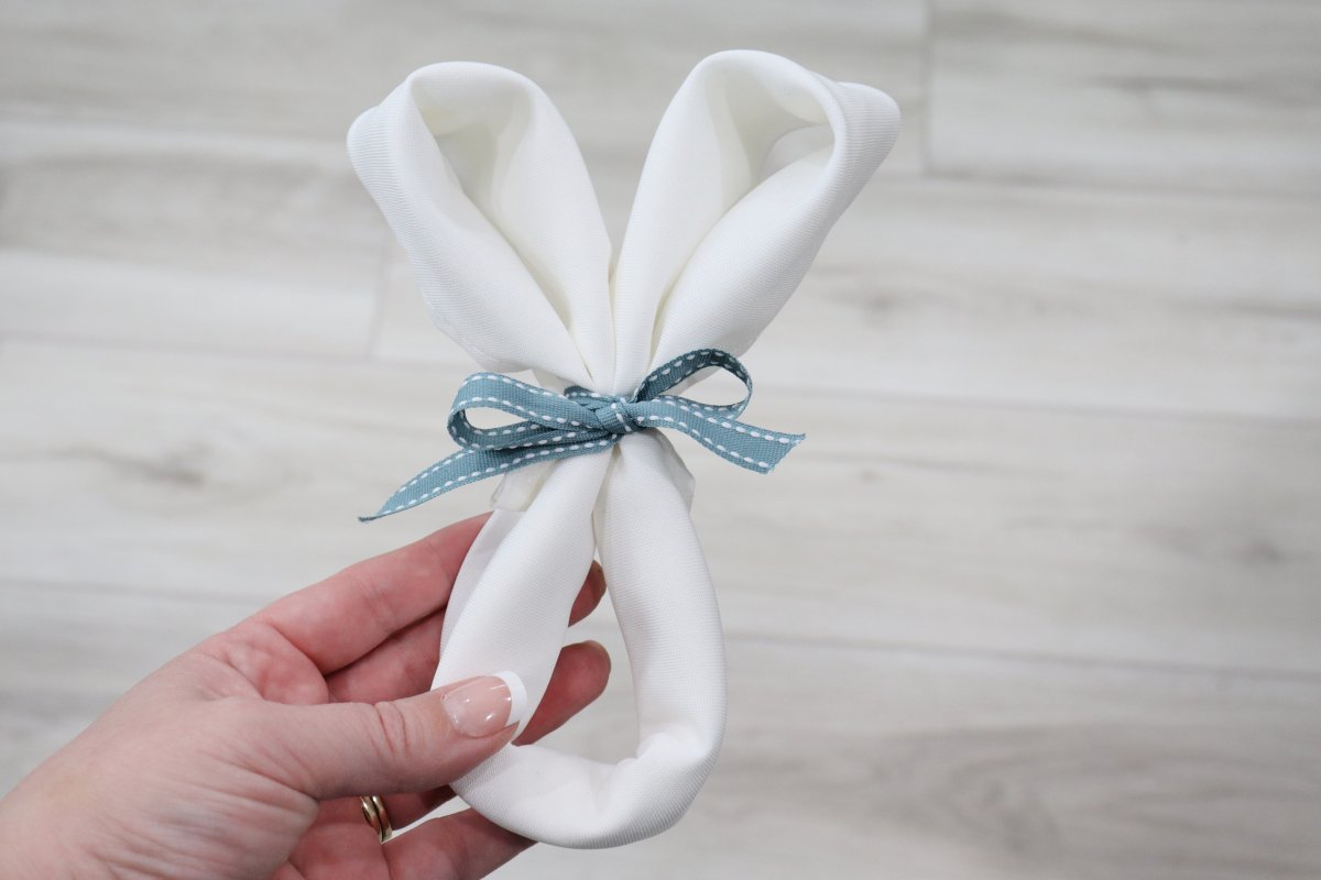 Image shows Amy's hand holding a folded white cloth napkin that is shaped like a bunny's head and tied with a blue bow below the ears.