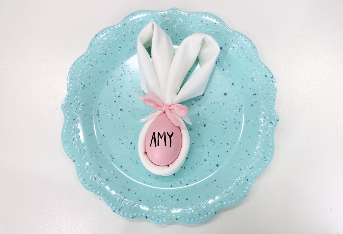 Image contains a white napkin folded in the shape of a bunny head and ears with a pink egg inside. The egg says, "Amy." It is sitting on a scalloped teal plate.