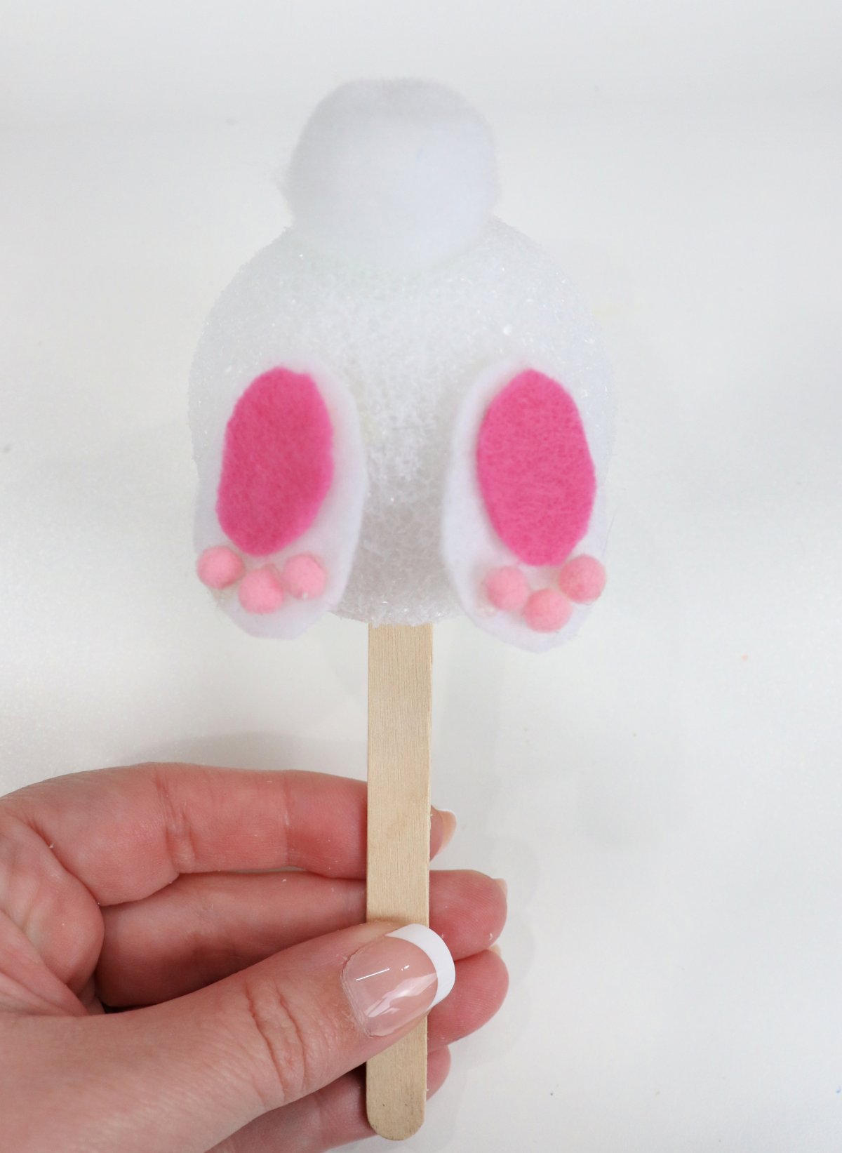 Image contains Amy's hand holding the bunny bottom made of a styrofoam ball with a white pom-pom tail, and pink and white felt feet, on a wooden craft stick.
