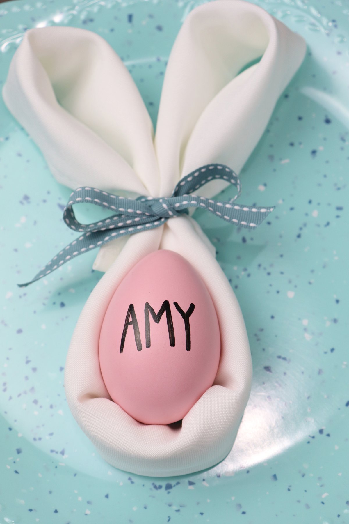 Image shows a white cloth napkin folded like a bunny head and ears with a blue bow. Inside the napkin is a pink egg with the name "Amy" written on it in black print. It sits on a teal plate.