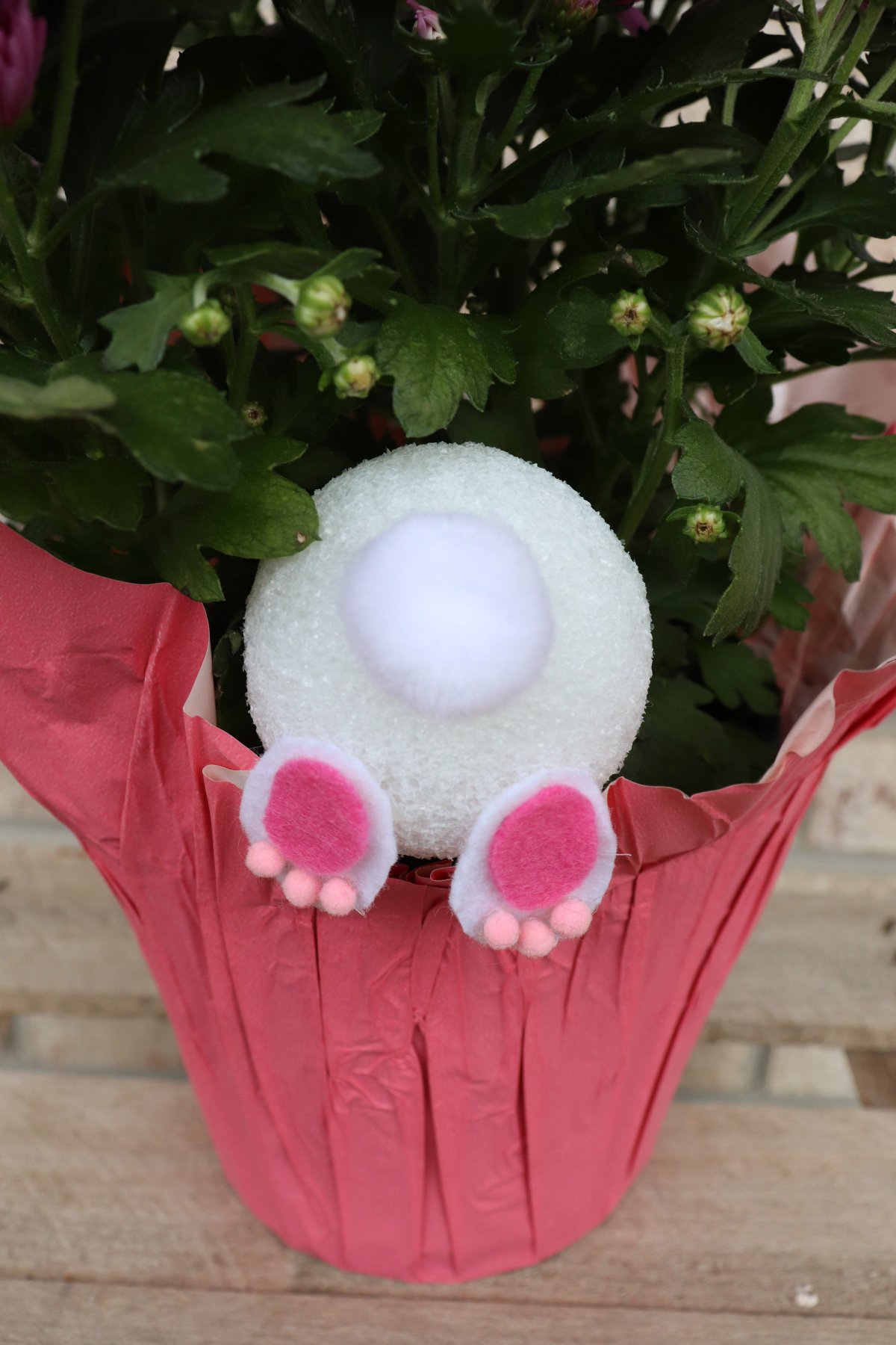 Image contains a potted purple mum plant with a "bunny bottom" made of a styrofoam ball, pom pom, and felt feet sticking out of the pot.