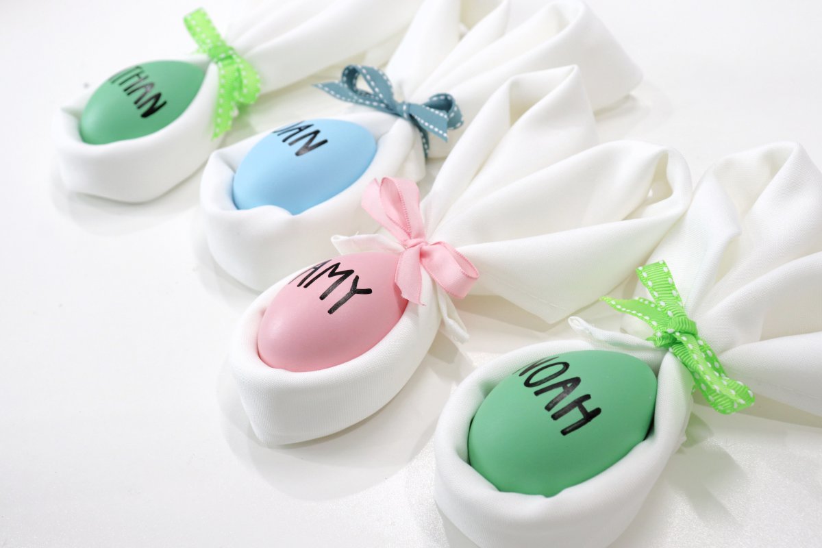 Image contains four folded white cloth napkins in the shape of a bunny with ears. Each one has a colored bow and a colored egg inside with a name printed on it. 