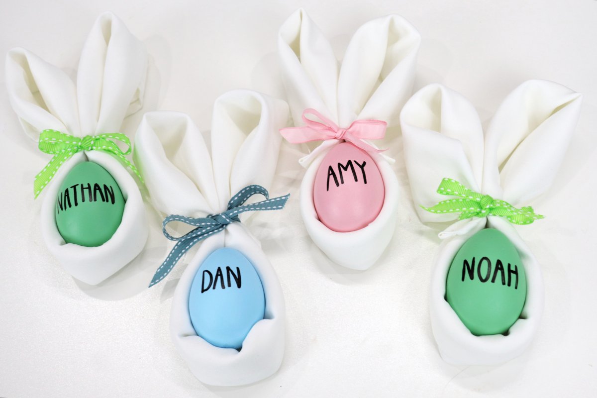 Image contains four folded white cloth napkins in the shape of bunnies. Each contains a colored egg with a name written on it in black marker.
