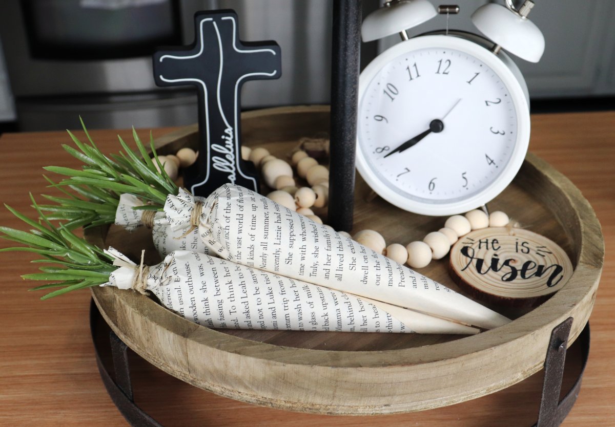 Image contains three carrots made from book pages displayed on a round wooden tray. The tray also contains a small black wooden cross, a natural wooden bead garland, and a white clock.