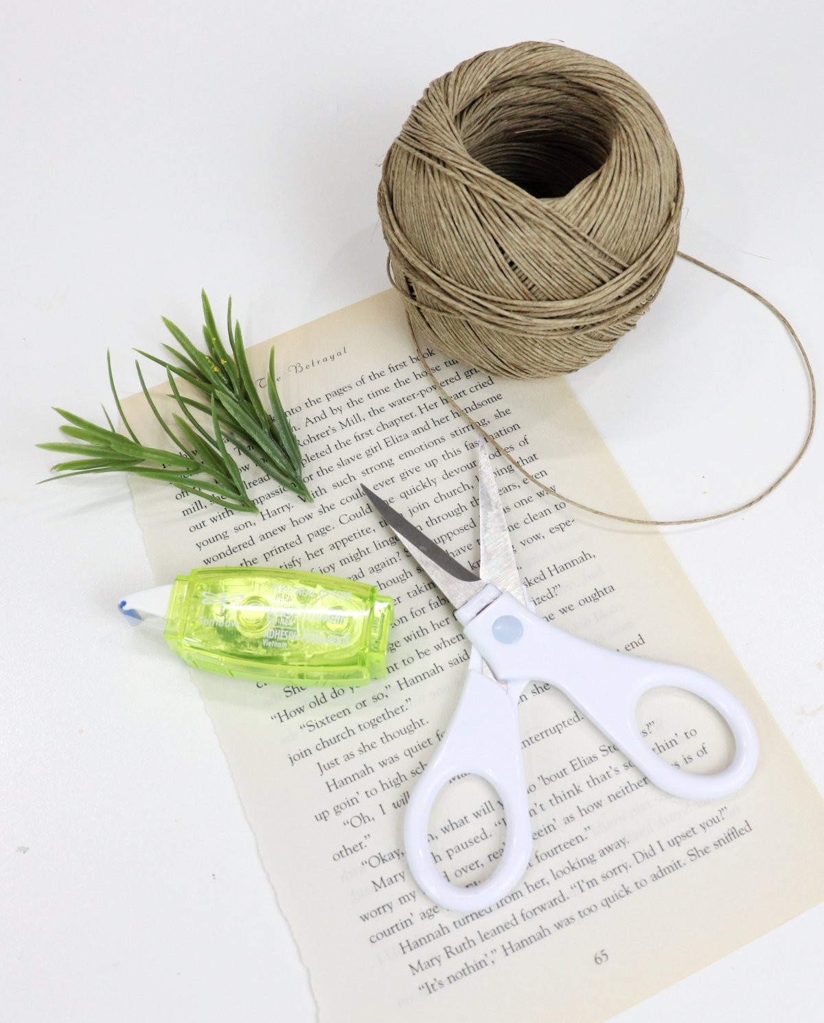 Image contains a book page, a white handled pair of scissors, a roll of adhesive, faux greenery, and a roll of jute on a white table.