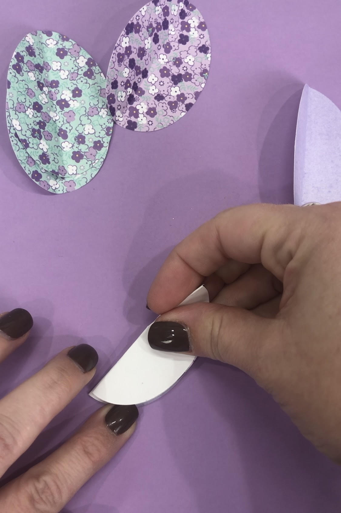 Image contains Amy's hands adhering a stack of folded egg shaped paper pieces together. It is on a purple background surrounded by egg shaped pieces of teal and purple patterned scrapbook paper.
